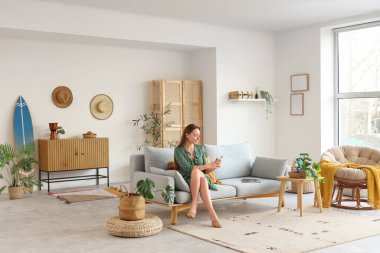 Oturma odasında sörf tahtasıyla koltukta cep telefonu kullanan genç bir kadın.