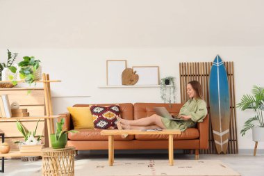 Oturma odasında sörf tahtasıyla kanepede dizüstü bilgisayar kullanan genç bir kadın.