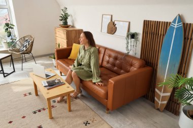 Oturma odasında sörf tahtasıyla kanepede oturan genç kadın.