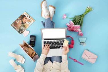 Dizüstü bilgisayarı ve kredi kartı olan bir kadın mavi arka planda online alışveriş yapıyor.