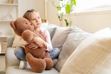 Evdeki kanepede oyuncak ayıya sarılan üzgün küçük kız.