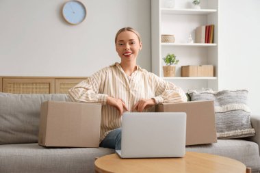 Paket kutuları ve dizüstü bilgisayarı olan genç bir kadın evdeki koltukta oturuyor.