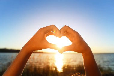 Kadın elleri gün batımında nehre karşı kalp hareketi yapıyor. Yaz gündönümü