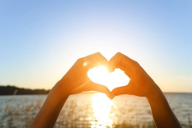 Kadın elleri gün batımında nehre karşı kalp hareketi yapıyor. Yaz gündönümü