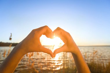 Kadın elleri gün batımında nehre karşı kalp hareketi yapıyor. Yaz gündönümü