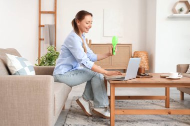 Mini fanı olan genç bir kadın evdeki koltukta dizüstü bilgisayar kullanıyor.