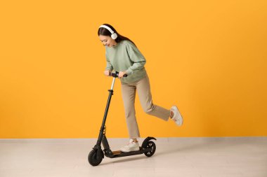 Kulaklıklı güzel bir Asyalı kadın sarı duvarın yanında modern elektrikli scooter kullanıyor.