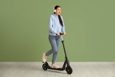 Yeşil duvarın yanında modern elektrikli scooterlı kulaklıklı güzel bir Asyalı kadın.