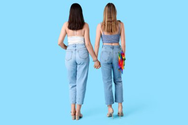 Mavi arka planda kot pantolon cebinde LGBT bayrağıyla el ele tutuşan genç lezbiyen çift.