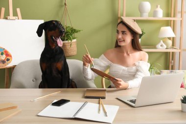 Sevimli Doberman köpeği ve evde elinde boya paleti olan şapkalı mutlu bir kadın.