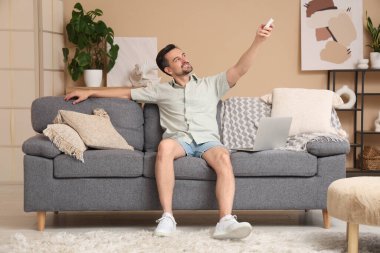 Genç adam oturma odasındaki bej duvarın yanındaki kanepede otururken klimayı açıyor.
