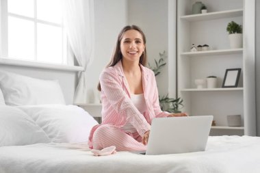 Pijamalı genç bir kadın evde dizüstü bilgisayar kullanıyor.