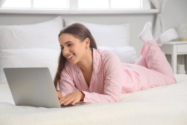 Pijamalı genç ve güzel bir kadın dizüstü bilgisayar kullanıyor ve evde yatakta yatıyor.