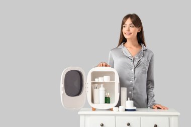 Gri arka planda kozmetik ürünleri için küçük buzdolabı olan güzel genç bir kadın.