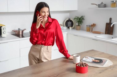 Güzel genç bir kadın cep telefonuyla konuşuyor ve mutfakta çikolata yiyor.