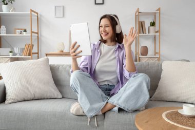 Kulaklıklı genç bir kadın evdeki kanepede tablet bilgisayar kullanıyor.