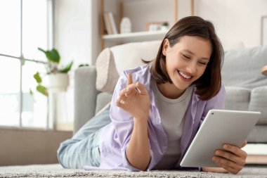 Evdeki halıda tablet bilgisayar kullanan genç bir kadın.