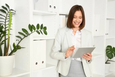 Ofiste rafların yanında tablet bilgisayarla çalışan genç bir iş kadını.