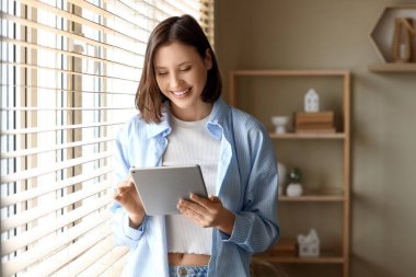 Genç bir kadın evdeki pencerenin yanında modern tablet bilgisayar kullanıyor.