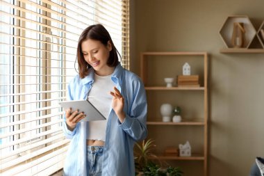 Genç bir kadın evdeki pencerenin yanında modern tablet bilgisayar kullanıyor.