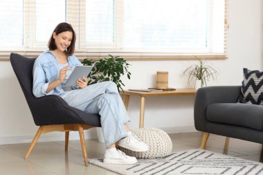 Evdeki koltukta tablet bilgisayar kullanan genç bir kadın.