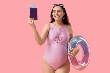 Pembe arka planda şişme yüzüğü ve pasaportu olan hamile bir kadın. Seyahat kavramı