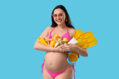 Mavi arka planda plaj aksesuarları olan hamile bir kadın. Seyahat kavramı