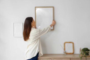 Young woman hanging blank frame on light wall at home, back view
