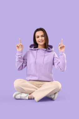 Burnunda kapüşonlu piercing olan genç bir kadın leylak arka plandaki bir şeyi işaret ediyor.