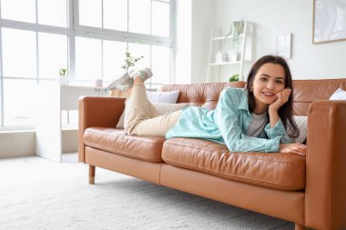 Burnunda piercing olan genç bir kadın evdeki kahverengi kanepede yatıyor.