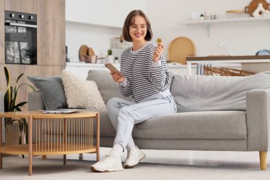 Evdeki koltukta oturan bitcoin ve cep telefonlu genç kadın.