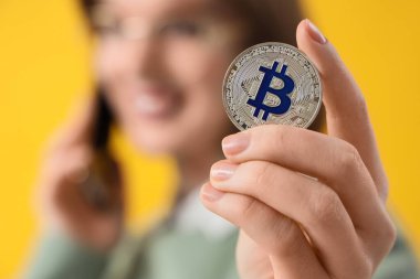 Bitcoin 'li genç kadın sarı arka planda cep telefonuyla konuşuyor, yakın plan.