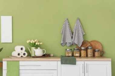 Çaydanlıklı makarna kavanozları, laleler ve kağıt havlular mutfaktaki yeşil duvarın yanındaki tezgahlarda.