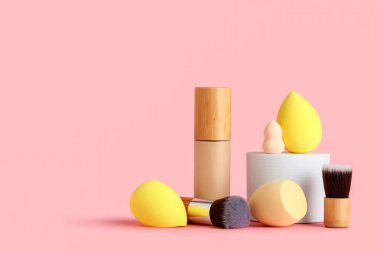 Pembe arka planda fırçalı makyaj süngeri ve bir şişe fondöten.