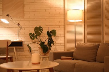 Gece oturma odasının içinde kanepe, katlanır ekran, parlayan lambalar ve beyaz tuğla duvarın yanındaki sehpa.