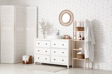 Banyo içinde, beyaz tuğla duvarın yanındaki çekmecelerin üzerinde vazoda çiçek açan dallar var.