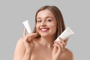 Yüzünde güneş kremi ve gri arka planda tüpler olan güzel genç bir kadın.