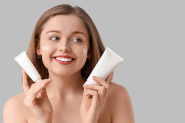 Yüzünde güneş kremi ve gri arka planda tüpler olan güzel genç bir kadın.