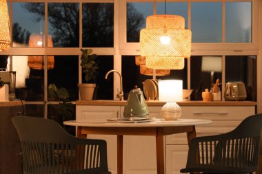 Geceleri mutfaktaki yemek masasında içinde dergiler ve parlayan lamba olan çaydanlık.