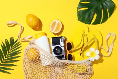 Sarı arka planda sicim torbası, plaj aksesuarları ve tropikal yapraklarla yaz kompozisyonu