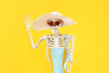 Sarı arka planda güneş gözlüğü ve yaz şapkası olan mayo giymiş insan iskeleti.