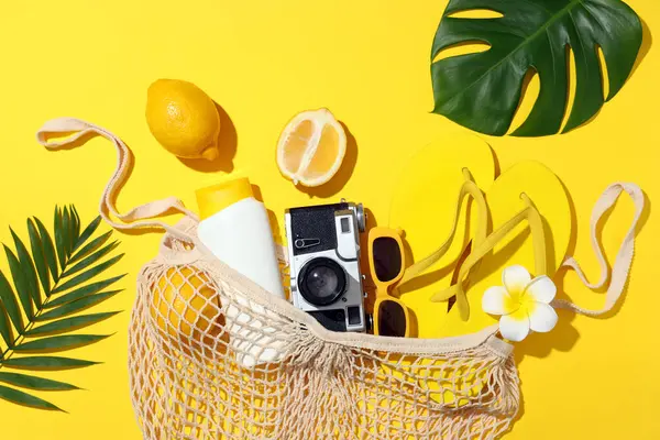 Sarı arka planda sicim torbası, plaj aksesuarları ve tropikal yapraklarla yaz kompozisyonu