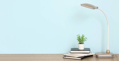 Çalışma odasındaki mavi duvarın yanında masa lambası, bitki ve defterler var. Tasarım için pankart