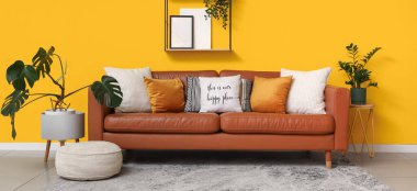 Yumuşak yastıklı kahverengi deri kanepe ve oturma odasındaki sarı duvarın yanında puf.