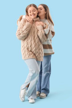 Sıcak süveter giyen genç lezbiyen çift mavi arka planda sarılıyorlar.
