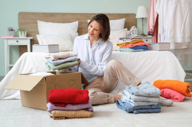 Genç ve güzel bir kadın farklı giysiler paketliyor. Evde yerde karton bir kutunun içinde.