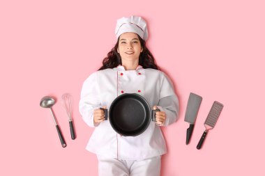 Farklı mutfak aletleri ve pembe arka planda duran çömleği olan mutlu genç bayan aşçı.