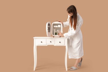 Bej arka planda kozmetik ürünleri için küçük buzdolabı olan güzel genç bir kadın.