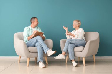 Yaşlı çift koltuklarda oturuyor ve mavi duvarın yanında konuşuyor.