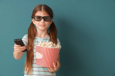 Üç boyutlu gözlüklü, patlamış mısırlı küçük kız mavi arka planda televizyon izliyor.